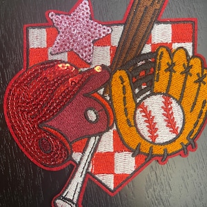May include: An embroidered patch featuring a red sequined baseball helmet, a brown baseball bat, a yellow baseball glove with a white baseball, and a pink sequined star, all set against a red and white checkered background with a red border.