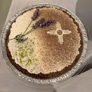 Può includere: Una torta rotonda in una teglia di alluminio argentato con una crosta marrone scuro. La parte superiore è divisa tra una farcitura color crema con scorza verde e un lato spolverato di marrone con un simbolo solare Zia bianco. Due rametti di lavanda.