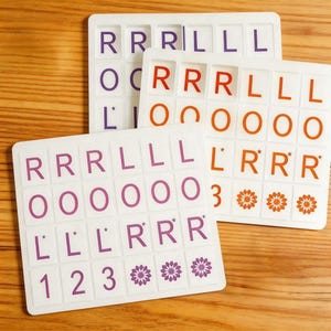 Könnte beinhalten: Drei weiße Spielbretter mit Buchstabenkacheln in Lila, Orange und Rot. Die Kacheln haben Buchstaben, Zahlen und Blumenmuster. Die Bretter liegen auf einer Holzoberfläche.