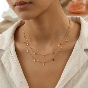 Op de afbeelding: Twee fijne, gouden kettingen met kleine vierkante bedels. De kettingen zijn gelaagd, waarbij de ene iets langer is dan de andere. De kettingen en bedels hebben een glanzende afwerking. De kettingen worden gedragen over een lichtgekleurd overhemd.