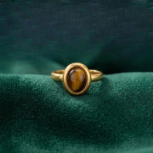 Peut inclure: Une bague dorée ornée d'une pierre précieuse œil-de-tigre ovale et lisse, sertie dans une monture métallique simple. La pierre présente des bandes chatoyantes brunes et dorées, tandis que l'anneau est en métal doré poli et uni.