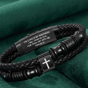 May include: A black braided leather bracelet featuring a rectangular metal plate engraved with the text THE [LORD] IS MY SHEPHERD: I WILL FEAR NO EVIL, FOR THOU ART WITH ME. PSALM 23. The bracelet includes a metal accent piece with a white cross.