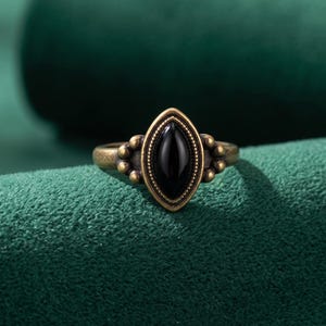 Peut inclure: Une bague dotée d'un anneau en métal couleur laiton antique avec une pierre noire centrale en forme de marquise. Le sertissage est encadré par une bordure perlée et agrémenté de petits détails métalliques sphériques de chaque côté.