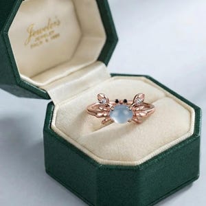 Peut inclure: Une bague en or rose en forme de crabe, dotée d'une pierre cabochon ronde bleu clair au centre pour le corps, de deux petites pierres sombres pour les yeux et de deux pierres claires incrustées dans les pinces, présentée dans un écrin hexagonal vert foncé.