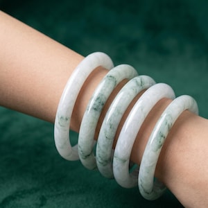 Peut inclure: Un ensemble de cinq bracelets joncs lisses et arrondis en jade blanc poli, agrémentés de subtiles inclusions vertes naturelles. Chaque bracelet présente une finition brillante et translucide ainsi qu'une forme circulaire uniforme.