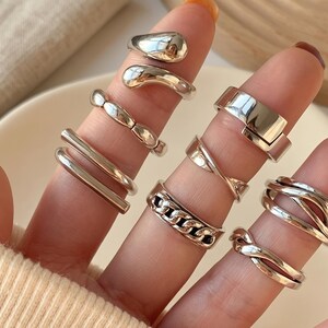 Peut inclure: Une collection de huit bagues en métal argenté portées sur les doigts. L'ensemble comprend divers designs tels que des anneaux lisses, un style chaîne, une bande torsadée, un anneau double et plusieurs formes organiques irrégulières ouvertes.