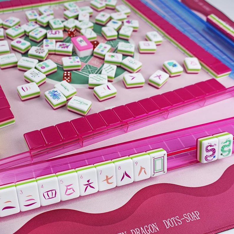 Puede incluir: Un juego de Mahjong rosa y blanco, con fichas dispersas sobre un tapete rosa. Las fichas son blancas con detalles verdes y rosas, que muestran caracteres y s&iacute;mbolos chinos. El juego incluye estantes y dados rosas.