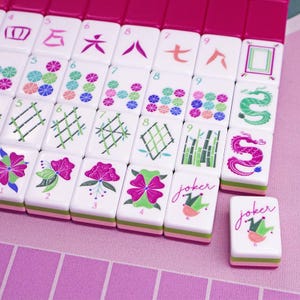 Puede incluir: Primer plano de un juego de Mahjong con fichas blancas que presentan varios s&iacute;mbolos, n&uacute;meros y dise&ntilde;os florales. Las fichas tienen detalles en rosa y verde, con un borde rosa. Dos fichas dicen "joker".