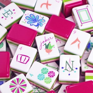 Puede incluir: Una colecci&oacute;n de coloridas fichas de Mahjong. Las fichas presentan varios dise&ntilde;os, incluyendo un pavo real, patrones florales y la palabra "Joker". Las fichas tienen una base blanca con acentos rosas y verdes.