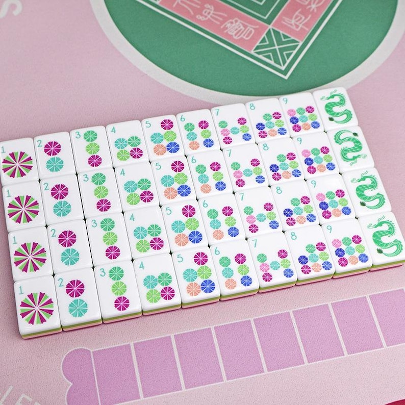 Puede incluir: Un juego de fichas de Mahjong blancas con dise&ntilde;os florales coloridos y n&uacute;meros. Las fichas est&aacute;n dispuestas sobre una superficie rosa con un fondo estampado en verde y rosa. El juego incluye fichas con ilustraciones de dragones verdes.