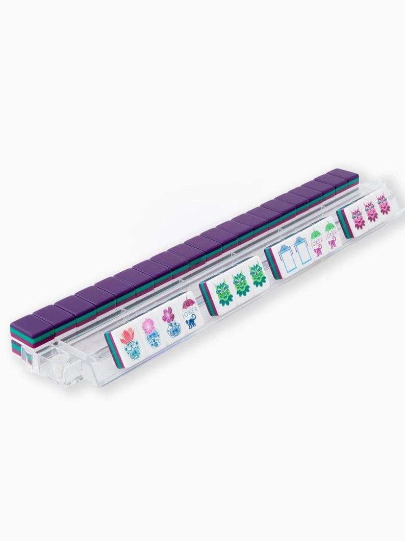 May include: A clear plastic tray holds a set of purple, teal, and white Mahjong tiles. The tiles are arranged in rows, with the purple tiles forming the majority. Some tiles have colorful floral and character designs.