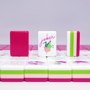 Puede incluir: Primer plano de fichas de Mahjong. Una ficha es magenta liso, otra tiene la palabra "Joker" con una ilustraci&oacute;n de p&aacute;jaro y corona, y la tercera tiene rayas verticales verdes, rosas y blancas. Las fichas est&aacute;n sobre un fondo blanco y rosa.
