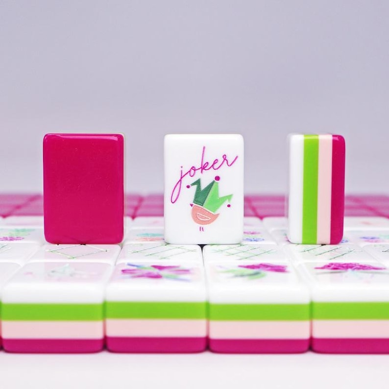 Puede incluir: Primer plano de fichas de Mahjong. Una ficha es magenta liso, otra tiene la palabra "Joker" con una ilustraci&oacute;n de p&aacute;jaro y corona, y la tercera tiene rayas verticales verdes, rosas y blancas. Las fichas est&aacute;n sobre un fondo blanco y rosa.