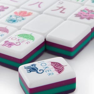May include: A set of white Mahjong tiles with colorful accents. Each tile features a white top, a purple middle, and a green base. Some tiles have illustrations of a monkey, an umbrella, and Chinese characters.
