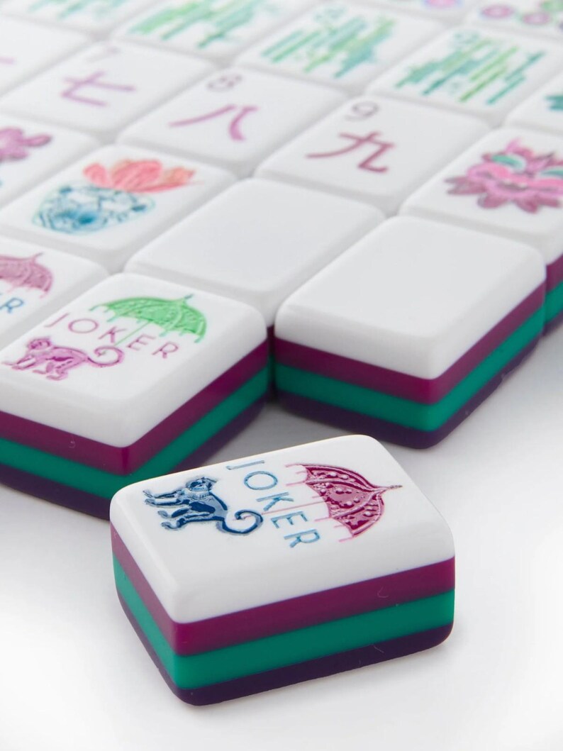 May include: A set of white Mahjong tiles with colorful accents. Each tile features a white top, a purple middle, and a green base. Some tiles have illustrations of a monkey, an umbrella, and Chinese characters.