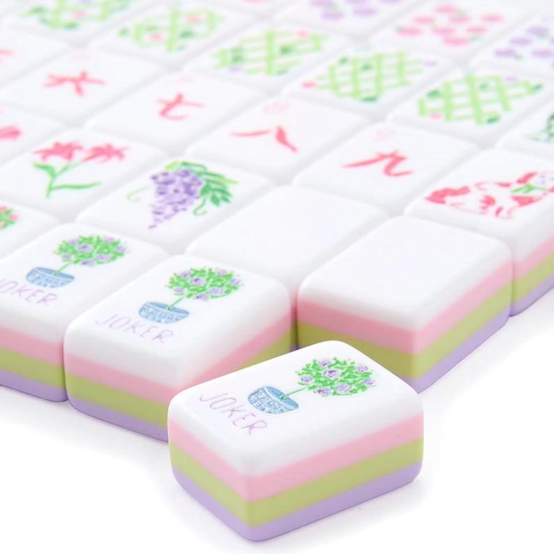 May include: A collection of white Mahjong tiles with colorful designs. The tiles feature illustrations of flowers, plants, and Chinese characters. The bottom of each tile has a layered design of pink, green, and purple. One tile reads "JOKER".