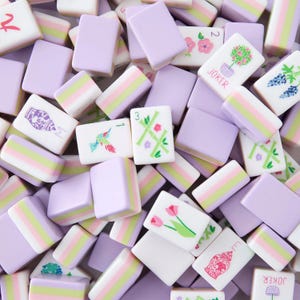 May include: Overhead shot of a collection of Mahjong tiles in shades of lavender, white, and pastel stripes. Some tiles feature floral and bird illustrations, while others display numbers and symbols. The word "JOKER" is visible on some tiles.