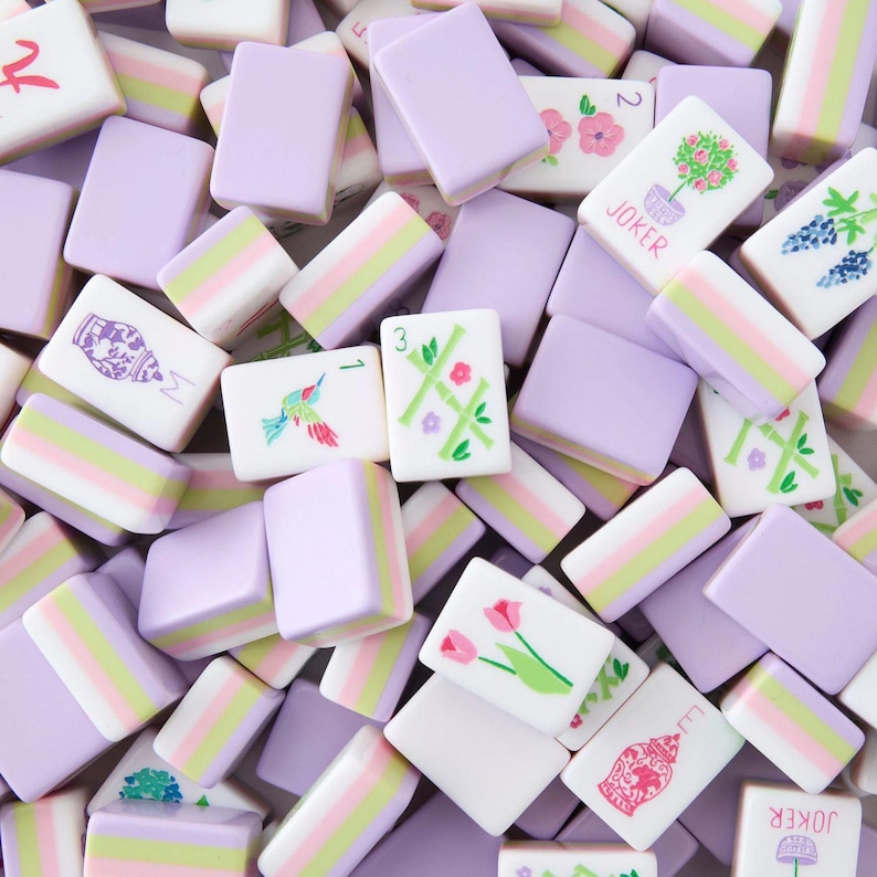 May include: Overhead shot of a collection of Mahjong tiles in shades of lavender, white, and pastel stripes. Some tiles feature floral and bird illustrations, while others display numbers and symbols. The word "JOKER" is visible on some tiles.