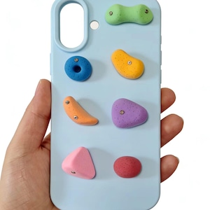 May include: A light blue phone case featuring colorful, textured climbing holds in various shapes and colors. The holds are in shades of blue, orange, yellow, purple, pink, and red. The case has a cutout for the camera and a hole for the charging port.