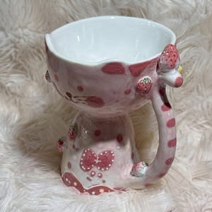 May include: A whimsical, pink ceramic mug with a white interior. The mug features a strawberry design, including a strawberry-shaped handle and decorative strawberries. The base has a bow design. The mug is approximately 4 inches tall.