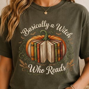 May include: A dark olive green t-shirt featuring a graphic of a pumpkin with open books inside. The text reads "Basically a Witch Who Reads" in a decorative font. The design includes small bats and wheat stalks surrounding the central pumpkin.