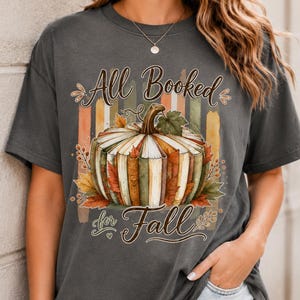 May include: A charcoal gray short-sleeve t-shirt featuring a graphic of a pumpkin made of stacked books. The text reads All Booked for Fall. The design includes vertical stripes in muted autumn colors and scattered fall leaves around the pumpkin.