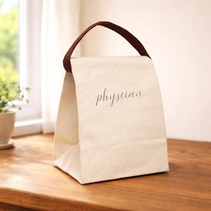 Może przedstawiać: Kremowa płócienna torba na lunch z brązową rączką. Słowo "physician" jest wydrukowane kursywą na przodzie. Torba stoi na drewnianej powierzchni.