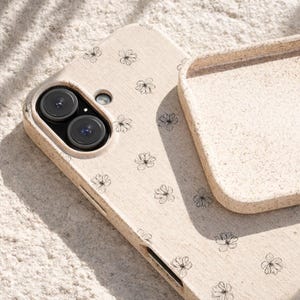 May include: A beige phone case with a floral pattern. The case has a dual-lens camera cutout and a separate top piece. The floral design consists of small, black, hand-drawn flowers scattered across the surface.