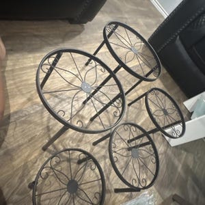 May include: Black metal plant stands with decorative circular tiers. The stands have a floral design in the center of each tier and are supported by three legs. The stands are designed to hold multiple plants.