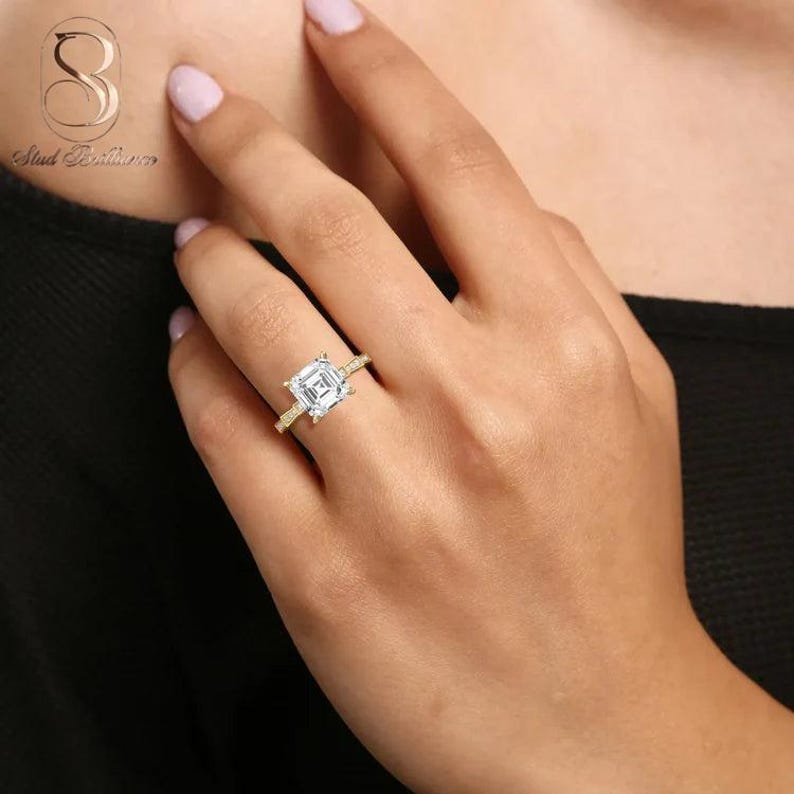 K&ouml;nnte beinhalten: Ein Goldring mit einem gro&szlig;en, quadratisch geschliffenen, klaren Mittelstein mit abgestuftem Facettenmuster. Das Band ist d&uuml;nn und metallisch und weist ein poliertes Gold-Finish auf, das die brillante, reflektierende Qualit&auml;t erg&auml;nzt.