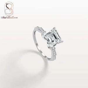 K&ouml;nnte beinhalten: Ein silberfarbener Ring mit einem gro&szlig;en, quadratisch geschliffenen, klaren Mittelstein, der von vier Krappen gehalten wird. Die Ringschiene ist mit kleinen, runden, klaren Steinen besetzt und das Logo Stud Brilliance ist oben links zu sehen.