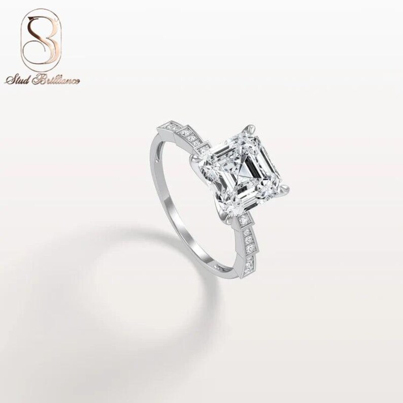 K&ouml;nnte beinhalten: Ein silberfarbener Ring mit einem gro&szlig;en, quadratisch geschliffenen, klaren Mittelstein, der von vier Krappen gehalten wird. Die Ringschiene ist mit kleinen, runden, klaren Steinen besetzt und das Logo Stud Brilliance ist oben links zu sehen.
