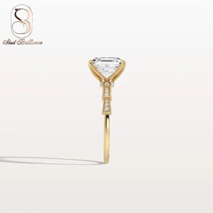K&ouml;nnte beinhalten: Ein Ring aus Gelbgold mit einem gro&szlig;en Smaragdschliff-Edelstein, der von vier Krappen gehalten wird. Die Ringschiene und die Galerie sind mit kleinen runden Pav&eacute;-Diamanten besetzt. Das Stud Brilliance Logo ist oben links im Bild zu sehen.