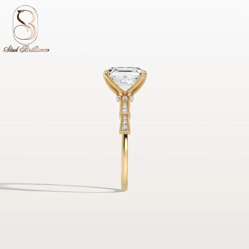 K&ouml;nnte beinhalten: Ein Ring aus Gelbgold mit einem gro&szlig;en Smaragdschliff-Edelstein, der von vier Krappen gehalten wird. Die Ringschiene und die Galerie sind mit kleinen runden Pav&eacute;-Diamanten besetzt. Das Stud Brilliance Logo ist oben links im Bild zu sehen.