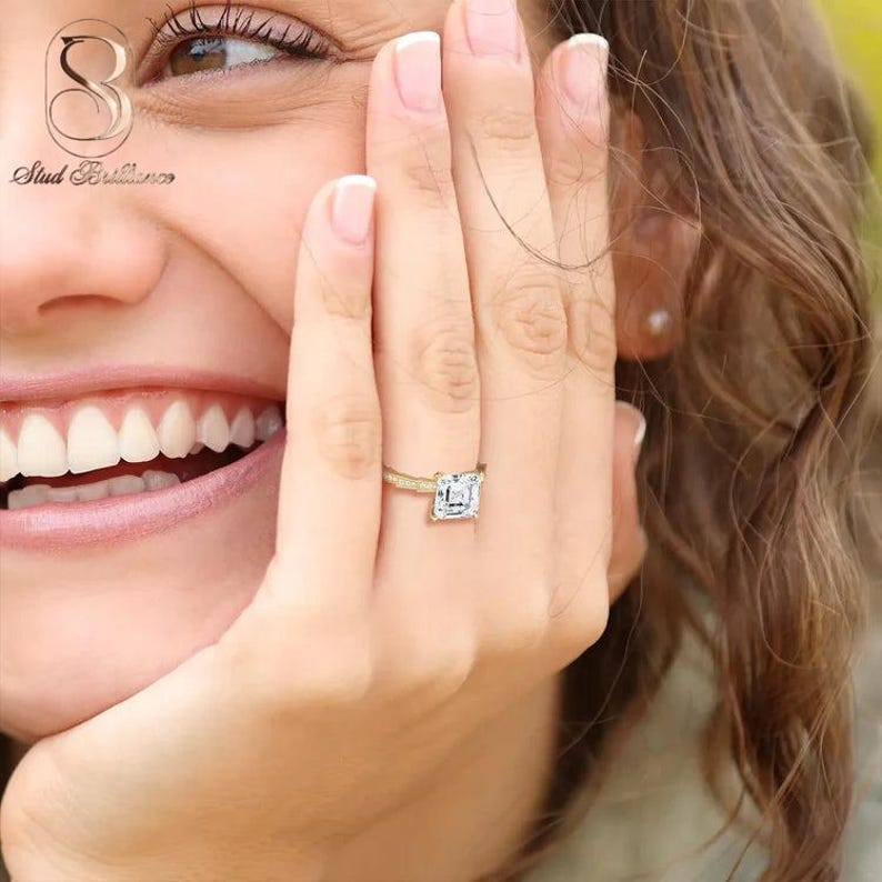 K&ouml;nnte beinhalten: Ein goldener Ring mit einem quadratisch geschliffenen, klaren Edelstein in der Mitte und einer d&uuml;nnen Ringschiene, die an den Seiten mit kleinen Pav&eacute;-gefassten Steinen besetzt ist. Der Ring sitzt an einem Finger und zeigt das Metall.