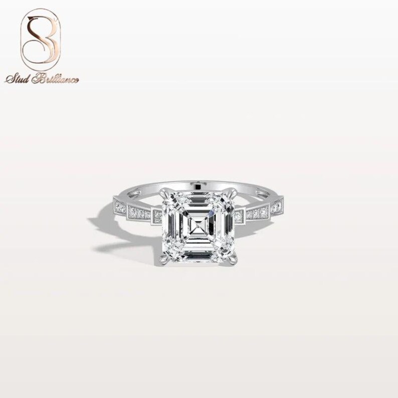 K&ouml;nnte beinhalten: Ein silberfarbener Ring mit einem gro&szlig;en, quadratisch geschliffenen Mittelstein mit Treppenschliff-Facettenmuster. Die Ringschiene hat eine metallische Oberfl&auml;che und ist mit kleinen, klaren, rund geschliffenen Steinen an den Seiten besetzt.