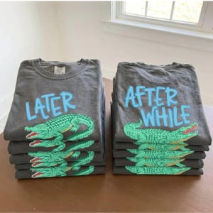 May include: Stacks of gray t-shirts with the phrases "LATER" and "AFTER WHILE" in light blue. Each shirt features a green alligator design with red accents. The shirts are neatly folded and stacked on a brown surface.