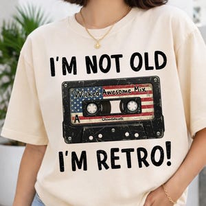 Può includere: T-shirt color crema con la scritta "I'M NOT OLD I'M RETRO!" sopra e sotto una grafica a cassetta. Il design della cassetta presenta la bandiera americana e le parole "Wicked Awesome Mix".