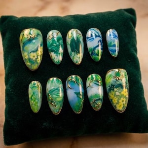 May include: A set of ten artificial nails with a nature-inspired design. Each nail features a base of green, blue, and yellow hues, with gold accents. The nails are arranged on a dark green velvet surface.