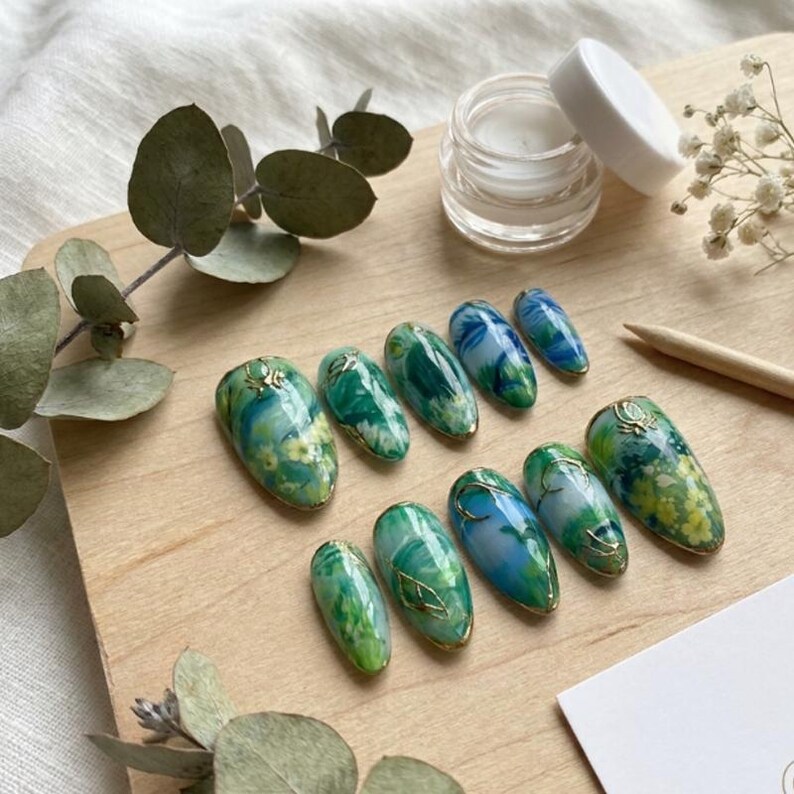 May include: Ten oval-shaped artificial nails with a marbled design in green and blue, with gold accents. A small jar of cream and sprigs of greenery are arranged on a wooden surface. The nails are approximately 2 cm in length.