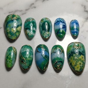 May include: A set of ten oval-shaped artificial nails. Each nail is decorated with a unique design in shades of green, blue, and yellow, with gold edging. The designs include floral and abstract patterns.