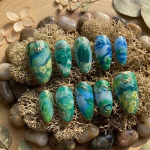 May include: A collection of ten oval-shaped false nails. Each nail showcases a unique design with green, blue, and yellow hues, complemented by gold detailing. The nails are displayed on a bed of moss and small stones, creating a natural aesthetic.