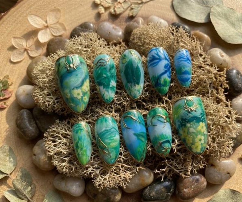 May include: A collection of ten oval-shaped false nails. Each nail showcases a unique design with green, blue, and yellow hues, complemented by gold detailing. The nails are displayed on a bed of moss and small stones, creating a natural aesthetic.