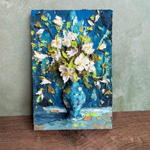May include: A textured painting of white flowers in a blue vase against a blue background. The artwork features a thick impasto technique, with visible brushstrokes and a three-dimensional effect. The flowers have yellow centers and green leaves.
