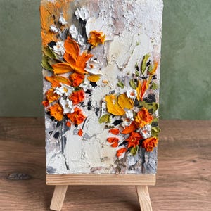 Könnte beinhalten: Ein kleines, strukturiertes Gemälde auf einer Holzstaffelei. Das Kunstwerk zeigt ein Blumendesign mit dicken Pinselstrichen in Orange-, Weiß-, Gelb- und Grüntönen vor einem strukturierten weißen und grauen Hintergrund. Die Staffelei ist aus hellem Holz.