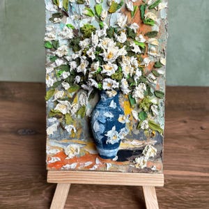 May include: A textured painting of white flowers in a blue vase with white floral accents. The vase sits on a wooden easel. The background features a mix of orange, green, and blue hues, creating a vibrant and artistic display.