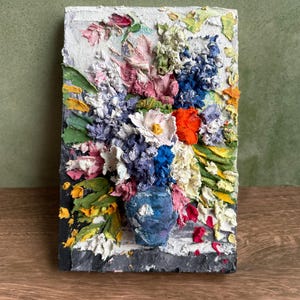 Puede incluir: Obra de arte texturizada de un arreglo floral en un jarrón azul. Las flores son una mezcla de blanco, rosa, azul y naranja, con hojas verdes. La obra está sobre una superficie rectangular texturizada.