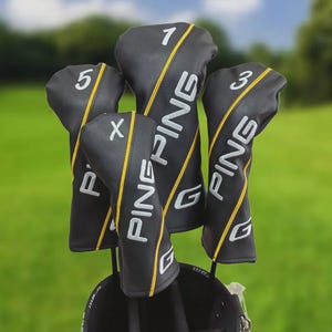 Puede incluir: Fundas negras para palos de golf con texto "PING" blanco y detalles amarillos. Cada funda está etiquetada con un número o letra: 1, 3, 5 y X. Las fundas están en una bolsa de golf sobre un fondo verde.