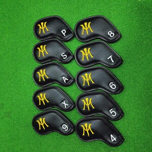 K&ouml;nnte beinhalten: Ein Satz von zehn schwarzen Golfschl&auml;gerhauben mit goldgestickten "M"-Logos. Jede Haube hat eine wei&szlig;e Nummer oder einen Buchstaben, der den Schl&auml;ger angibt. Die Hauben sind auf einer gr&uuml;nen Oberfl&auml;che angeordnet.