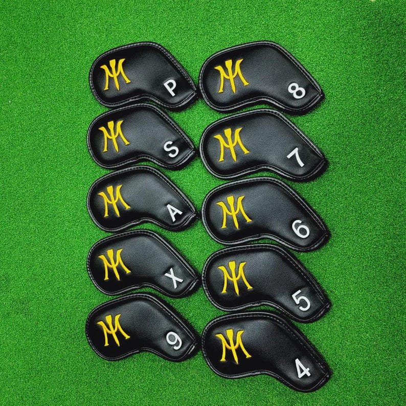 K&ouml;nnte beinhalten: Ein Satz von zehn schwarzen Golfschl&auml;gerhauben mit goldgestickten "M"-Logos. Jede Haube hat eine wei&szlig;e Nummer oder einen Buchstaben, der den Schl&auml;ger angibt. Die Hauben sind auf einer gr&uuml;nen Oberfl&auml;che angeordnet.